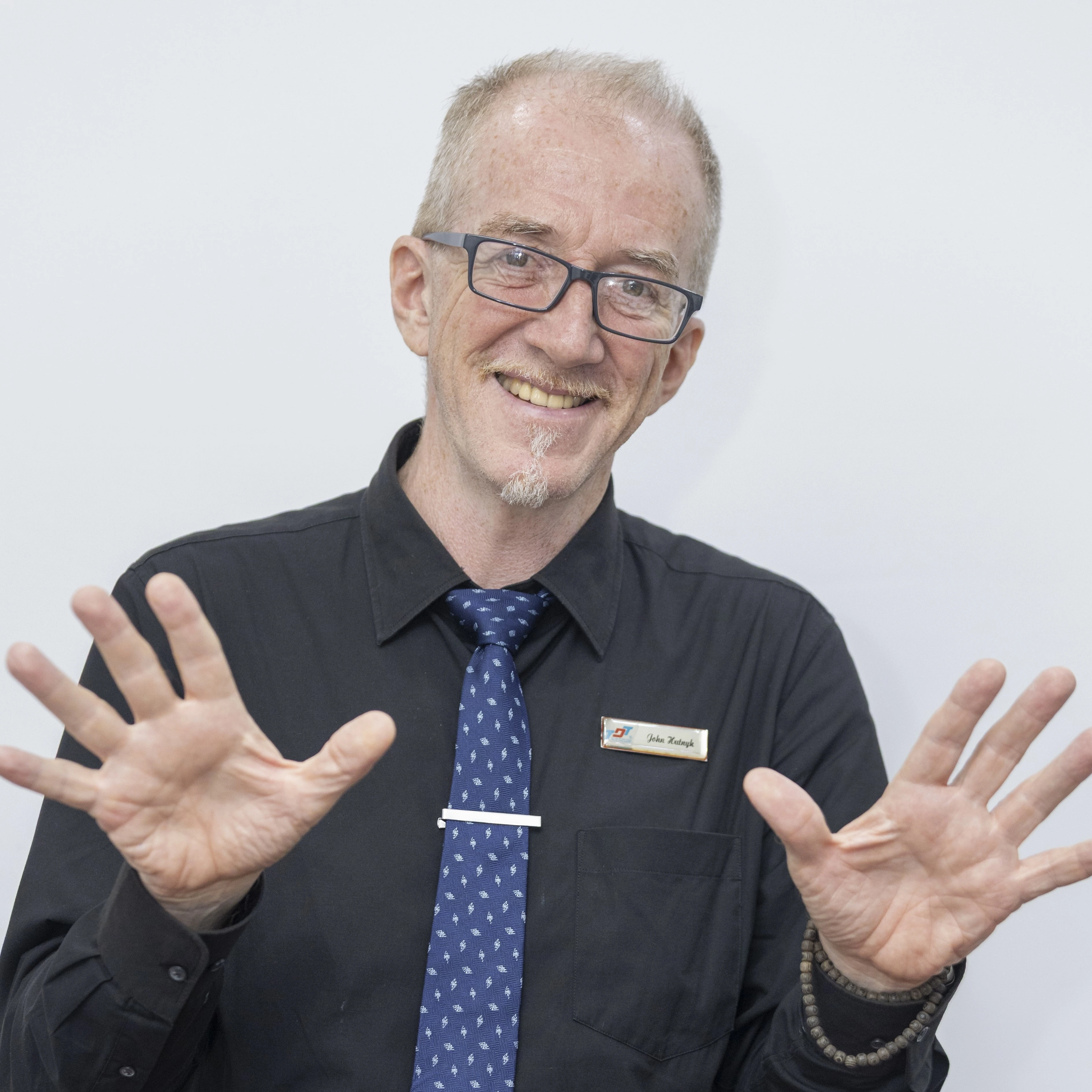 A smiling older man wearing glasses, a black shirt, and a blue patterned tie raises both hands with open palms. He has short gray hair and a small goatee, and a name badge is pinned to his shirt. The background is plain and light-colored.