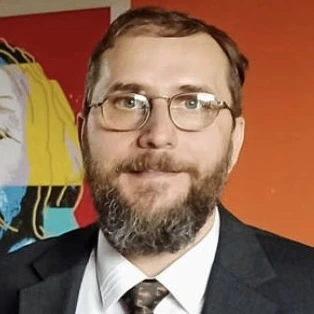 A man with glasses and a full beard looks directly at the camera, wearing a dark suit, white shirt, and patterned tie. The background features a colorful wall with abstract artwork.